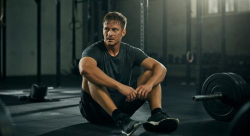 Erschöpfte Athletin sitzt auf dem Gym-Boden als Sinnbild für starken Muskelkater nach einem WOD.